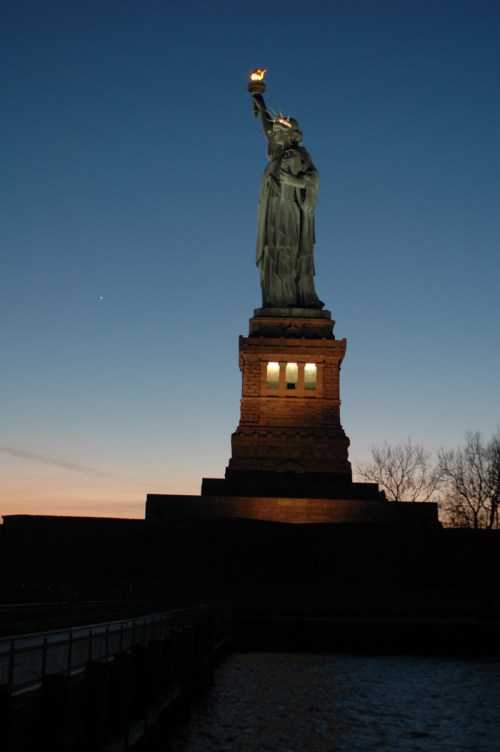 The Statue of Liberty stands tall against a twilight sky, holding a torch, with silhouettes of trees in the foreground.
