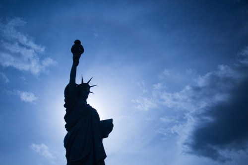 Silhouette of the Statue of Liberty against a blue sky with clouds, holding a torch.