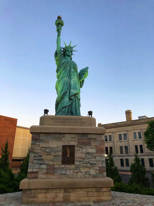 Statue of Liberty replica standing tall against a clear blue sky, with greenery and buildings in the background.