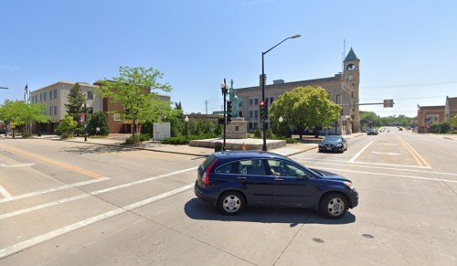 A blue car turns at an intersection near a clock tower and green trees on a sunny day.