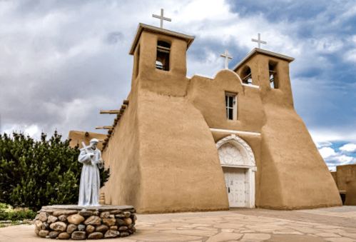 Historic adobe church with two bell towers and a statue in front, set against a cloudy sky.