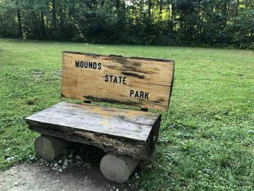 Wooden bench with "Mounds State Park" carved on the back, set in a grassy area surrounded by trees.