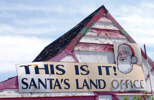 Sign on a building reads "THIS IS IT! SANTA'S LAND OFFICE" with a Santa illustration, under a blue sky.