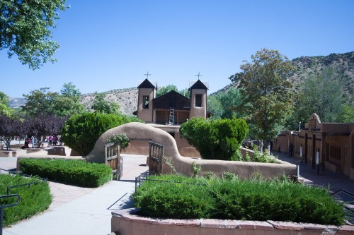 A scenic view of a historic adobe church surrounded by greenery and trees under a clear blue sky.