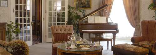 Elegant parlor with a grand piano, plush seating, and a table set with tea and pastries, surrounded by plants.