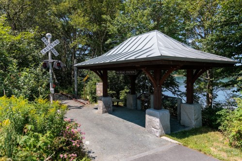 A wooden shelter at a train station near a crossing, surrounded by greenery and overlooking a body of water.