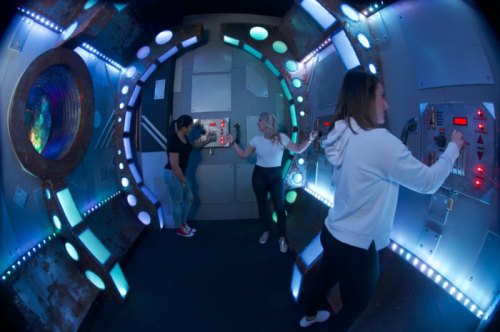 Three people interact with futuristic control panels in a colorful, sci-fi themed room with glowing lights.