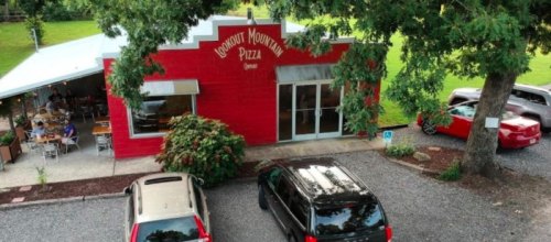 A red pizza restaurant with outdoor seating, surrounded by trees and parked cars in a gravel lot.