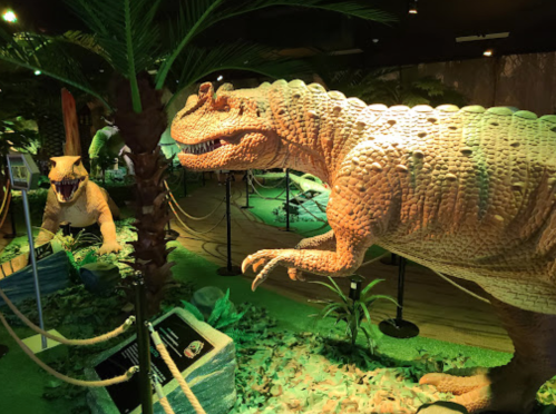 Two large dinosaur models in a lush, green exhibit with palm trees and informational signs.