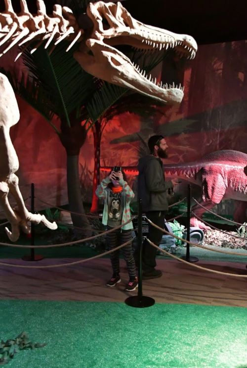 A child and an adult explore a dinosaur exhibit with large dinosaur models in a dimly lit environment.