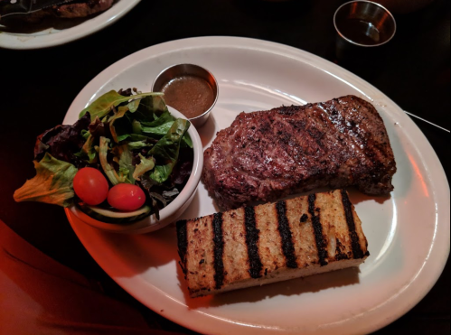 A plate with a grilled steak, a side salad with cherry tomatoes, and a slice of grilled bread.