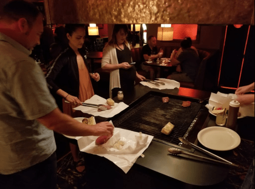A group of people cooking meat on a grill at a restaurant, with plates and condiments on the table.