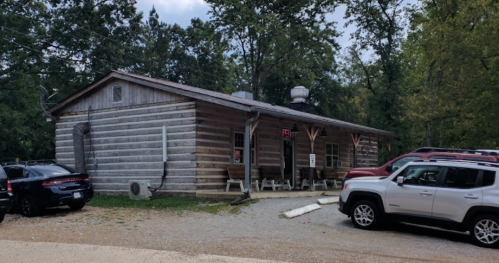 A rustic log cabin building with a gravel parking area, surrounded by trees and parked cars.