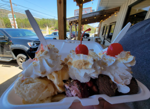 A takeout container filled with ice cream, whipped cream, and cherries, held outdoors near a restaurant.