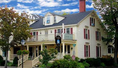 A charming yellow inn with red shutters, a porch, and a sign, set against a blue sky with scattered clouds.