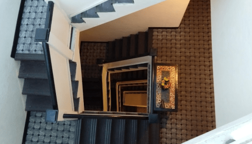 A top-down view of a staircase with dark steps and a patterned carpet, featuring a small table with flowers at the bottom.