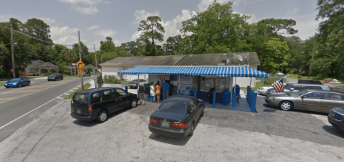 A small roadside eatery with blue awnings, surrounded by parked cars and trees, with people outside.