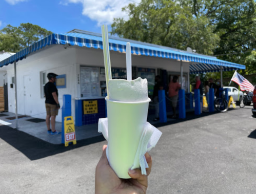 A hand holds a milkshake in front of a blue-striped ice cream stand with customers and an American flag nearby.