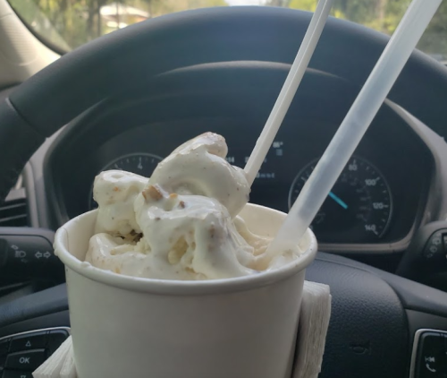 A cup of ice cream with two spoons, resting on a car's steering wheel with a blurred dashboard in the background.