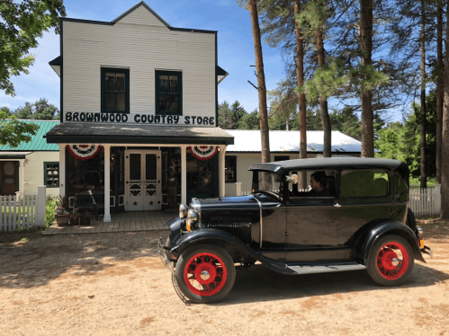 A vintage black car with red wheels parked in front of a country store surrounded by trees.