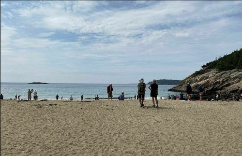A sandy beach with people walking and playing near the water, surrounded by rocky cliffs and a cloudy sky.