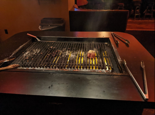 A smoky grill with a piece of meat cooking, surrounded by utensils on a dark table.
