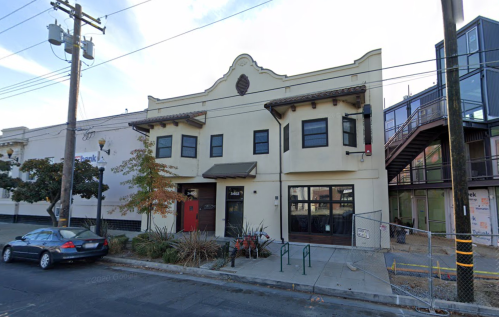 A two-story building with a decorative facade, featuring large windows and a red door, surrounded by plants and power lines.