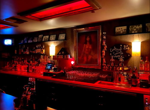 A dimly lit bar with red lighting, shelves of liquor, and a large portrait on the wall.