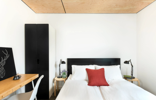A modern bedroom featuring a double bed, a red accent pillow, a desk, and a wardrobe against a minimalist backdrop.