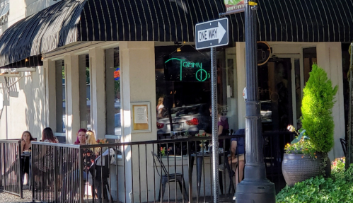 A corner restaurant with outdoor seating, featuring a striped awning and a neon sign, surrounded by greenery.