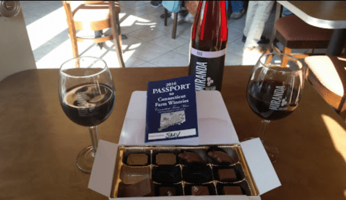 A table setting with two glasses of red wine, a bottle, a chocolate box, and a Connecticut Farm Wineries passport.