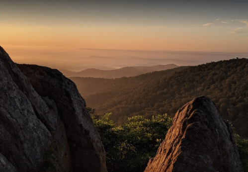 Sunset over rolling mountains, with rocky foreground and a hazy horizon, creating a serene and tranquil landscape.