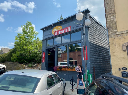 A diner with a gray exterior and large windows, featuring a sign that reads "Nick's Diner," surrounded by trees and cars.
