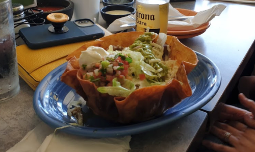 A colorful taco salad in a crispy shell, topped with lettuce, tomatoes, sour cream, and guacamole on a blue plate.