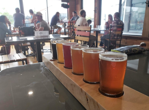 A flight of colorful craft beers on a wooden paddle, with people socializing in the background of a brewery.