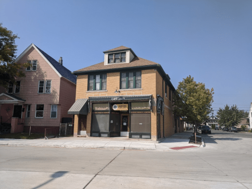 A brick building with a cafe on the ground floor, surrounded by trees and residential homes. Clear blue sky above.