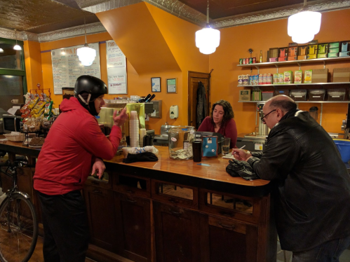 A coffee shop scene with a customer in a helmet talking to a barista, while another customer sits at the counter.