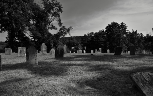 A black and white image of a cemetery with gravestones scattered across a grassy area under a cloudy sky.