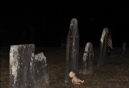 A dark graveyard scene with old gravestones and a doll lying on the ground, near an American flag in the background.