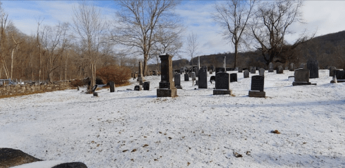 A snowy graveyard with various tombstones and bare trees under a cloudy sky.