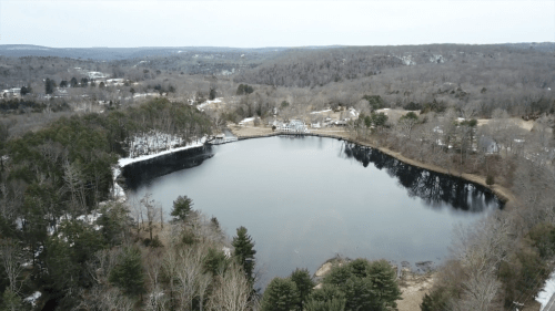 Aerial view of a serene lake surrounded by trees and hills, with a hint of snow on the ground.
