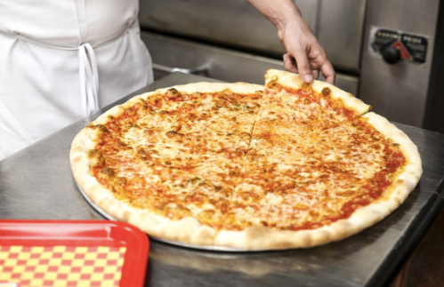 A person lifts a slice from a large cheese pizza on a metal tray in a kitchen setting.