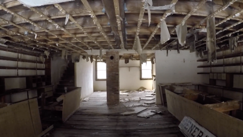 An abandoned room with a damaged ceiling, exposed beams, and debris scattered on the floor. Light enters through windows.
