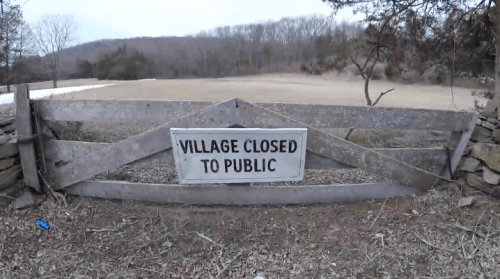 Sign reading "Village Closed to Public" in a rural area with trees and a field in the background.