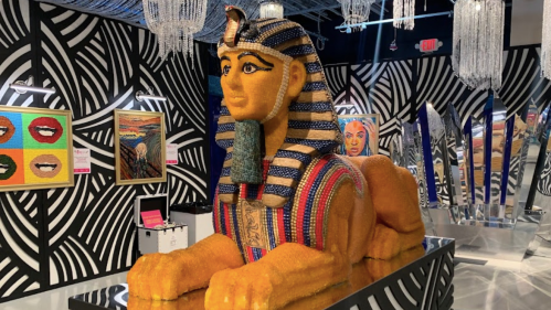 A colorful, large sphinx sculpture made of beads, displayed in a modern art gallery with vibrant artwork in the background.