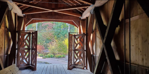 View from inside a wooden covered bridge, showcasing open doors and a glimpse of colorful foliage outside.