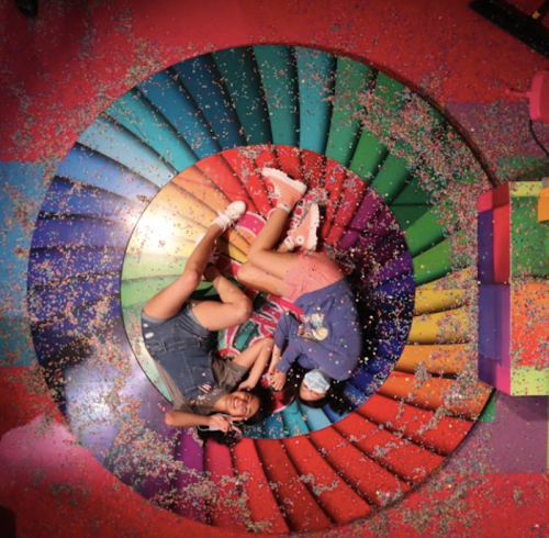 Two people lie on a colorful spiral floor covered in confetti, smiling and enjoying a playful moment together.