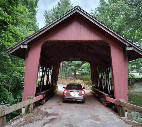 A red covered bridge with a car driving through, surrounded by lush green trees.