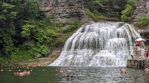 A scenic waterfall cascades into a swimming area, with people enjoying the water surrounded by lush greenery.