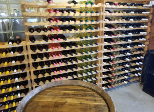 A wooden wine rack filled with various bottles, featuring colorful caps, beside a wooden barrel table.